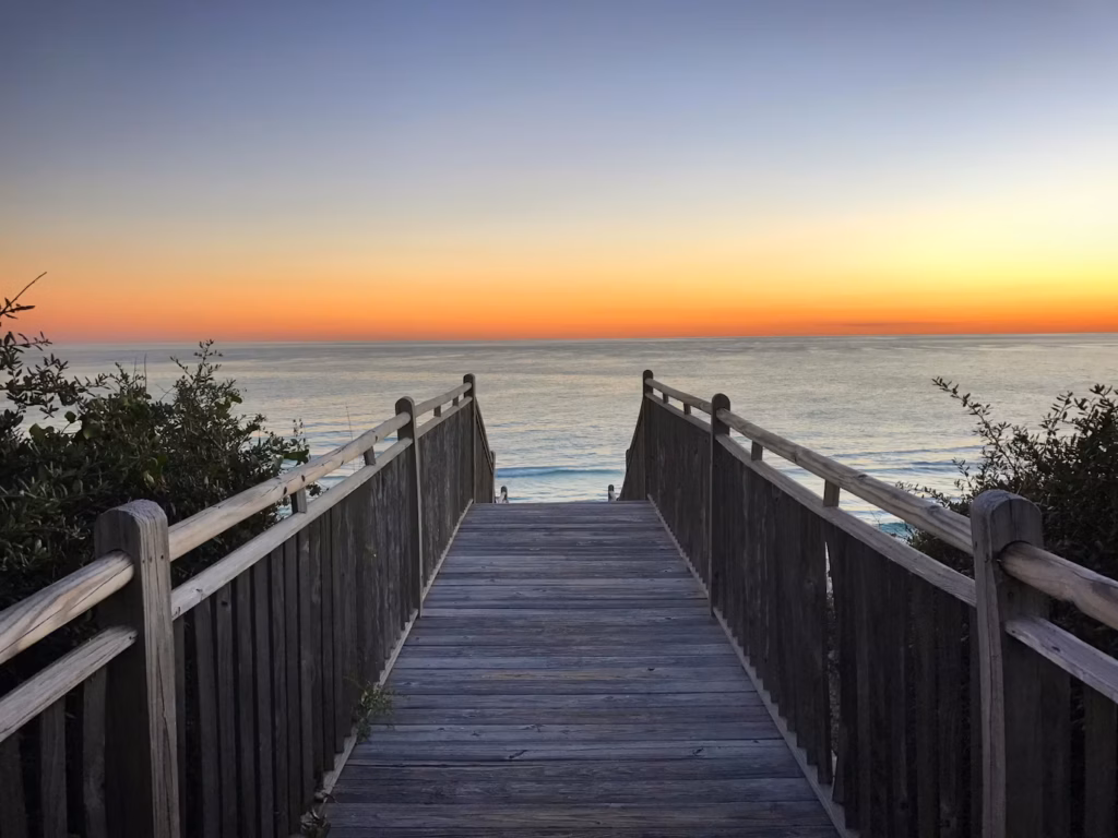A wooden boardwalk stretches toward a breathtaking sunset over Alys beach's waters.