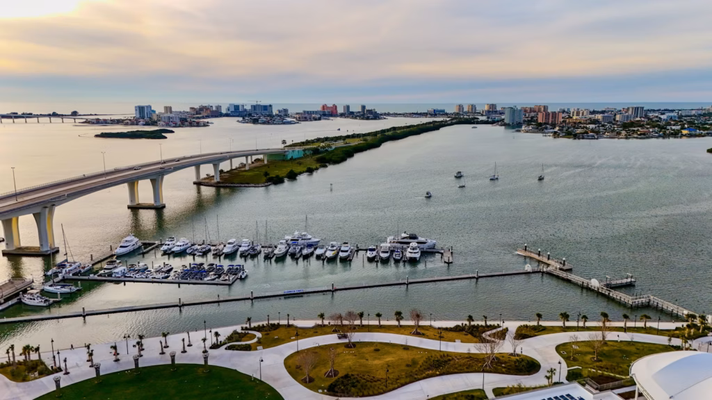 A prominent high bridge spans across the water in the foreground, connecting landmasses, with a modern marina full of luxury yachts and boats docked along wooden piers.