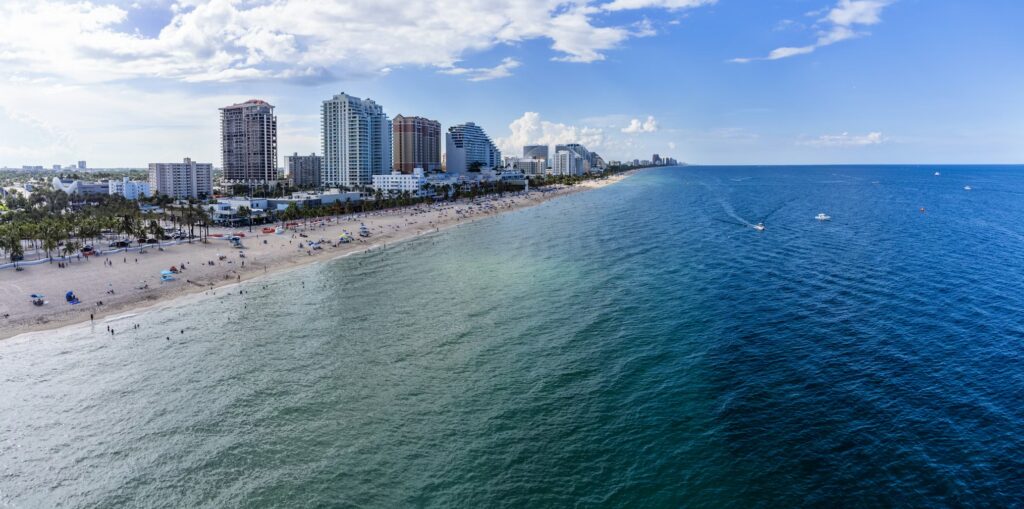 A breathtaking aerial view of Fort Lauderdale Beach in Florida, showcasing the miles of golden sandy shoreline lined with colorful umbrellas and beachgoers, and condominiums under a bright blue sky with scattered clouds. The turquoise Atlantic Ocean sparkles in the foreground, with gentle waves meeting the coast and a few boats cruising offshore.