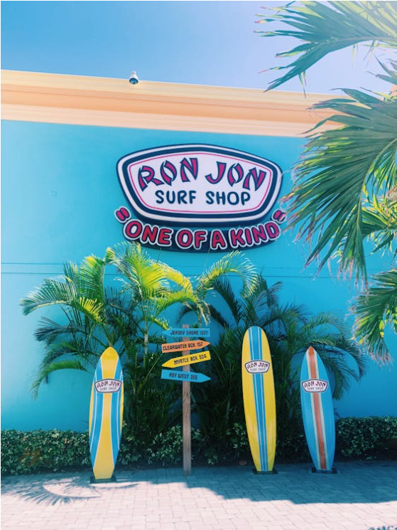 The bright turquoise wall serves as the backdrop for the large, colorful Ron Jon Surf Shop logo in white with red and blue accents, proudly displaying the slogan "ONE OF A KIND" in playful lettering. Three tall, classic surfboards painted in bright yellow, blue, and orange stand upright in front of the shop, each bearing the Ron Jon logo.